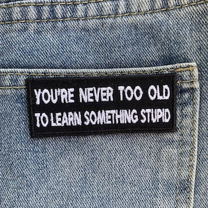 Könnte beinhalten: Nahaufnahme einer Jeans-Hosentasche mit einem schwarzen, gestickten Aufnäher. Der Aufnäher trägt die weiße Aufschrift "YOU'RE NEVER TOO OLD TO LEARN SOMETHING STUPID". Die Jeans hat eine hellblaue Waschung mit sichtbaren Nähten.