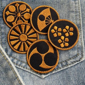 May include: Five embroidered patches with Japanese-inspired designs in gold and black. The patches feature various circular motifs, including a fan, flowers, and geometric patterns. The patches are displayed on a denim background.