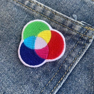 Pode incluir: Um remendo bordado colorido com um design de círculos sobrepostos em tons de verde, azul, amarelo, rosa e vermelho. O remendo é contornado de branco e tem um fundo branco.