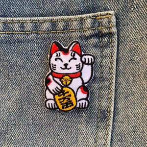 May include: An embroidered lucky cat patch in white and red, with a gold coin in its paw. The cat is smiling and has a bell around its neck. The patch is on a blue denim background.