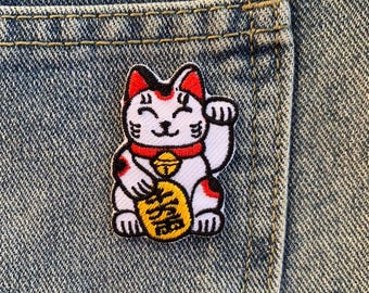 Peut inclure: Un patch brodé de chat porte-bonheur blanc et rouge avec une pièce d'or dans sa patte. Le chat sourit et a une cloche autour du cou. Le patch est sur un fond en jean bleu.