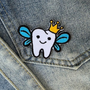 Puede incluir: Parche de diente blanco con una cara sonriente, alas azules y una corona dorada. El parche está delineado en negro y está sobre un fondo de mezclilla.