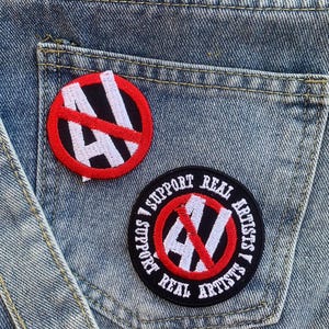 Puede incluir: Dos parches bordados sobre un fondo de mezclilla azul. El primer parche es rojo con una barra blanca que atraviesa la letra "A". El segundo parche es negro con texto blanco que dice "I Support Real Artists I Support Real Artists". El parche tiene un círculo rojo con una barra blanca que atraviesa la letra "A".