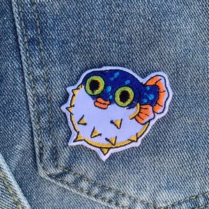 Puede incluir: Un parche bordado colorido con un pez globo de dibujos animados. El parche tiene un cuerpo azul con púas blancas, aletas naranjas y ojos verdes grandes. El parche está cosido en un bolsillo de chaqueta vaquera.