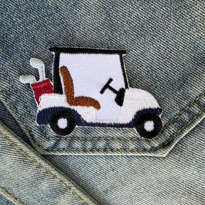 Op de afbeelding: Geborduurde patch van een golfkarretje met een witte carrosserie, zwarte wielen en een bruine stoel. De golfkar heeft een zwart stuur en een rode golftas met witte golfclubs. De patch staat op een denim achtergrond.