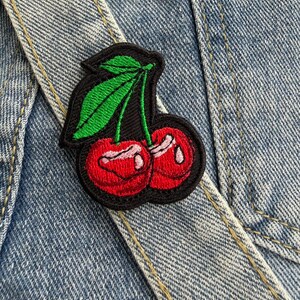 May include: Embroidered cherry patch featuring red cherries, green leaves, and a black border. The patch is attached to a denim jacket, highlighting the design's detail. The patch adds a splash of colour and a playful element to the denim.
