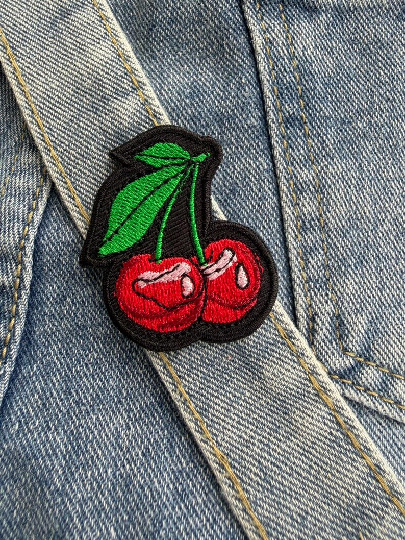 May include: Embroidered cherry patch featuring red cherries, green leaves, and a black border. The patch is attached to a denim jacket, highlighting the design's detail. The patch adds a splash of colour and a playful element to the denim.