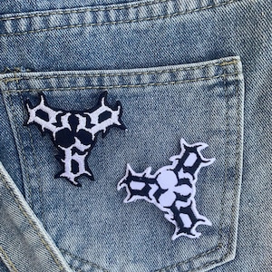 Puede incluir: Dos parches termoadhesivos en blanco y negro con forma triangular y diseño de calavera. Los parches están sobre un fondo de mezclilla azul.