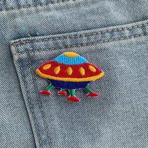 May include: A red, blue, and yellow embroidered UFO patch on a blue denim background.