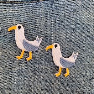 Puede incluir: Dos parches bordados para planchar de gaviotas sobre un fondo de mezclilla azul. Las gaviotas son blancas con alas grises y picos y patas naranjas.