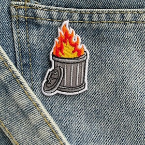 May include: Embroidered patch featuring a gray trash can with flames. The flames are depicted in shades of red, orange, and yellow. The patch is sewn onto a denim pocket, showcasing the patch's placement on clothing.