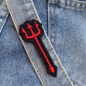 May include: Red and black embroidered trident patch on a blue denim background.