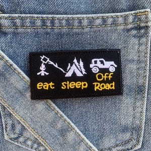 以下が含まれることがあります： キャンプファイヤー、テント、オフロード車を描いた黒と黄色の刺繍パッチ。テキストは「eat sleep off road」です。