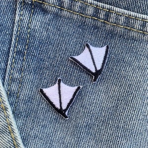 Puede incluir: Dos pequeños parches blancos con forma de pata de pato con contornos negros están adheridos a una tela vaquera azul. Los parches están bordados con costuras detalladas, y la mezclilla tiene costuras visibles y un aspecto texturizado.