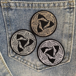 May include: Three embroidered iron-on patches with a black background and white, silver, and gold thread designs of three stylized birds in a circle.