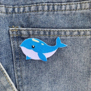 Könnte beinhalten: Ein blau-weißer Wal-Aufnäher ist auf der Gesäßtasche einer blauen Jeans aufgenäht. Der Wal hat einen hellblauen Körper, einen weißen Bauch und ein kleines schwarzes Auge. Die Jeans hat sichtbare Nähte und eine klassische Denim-Textur.