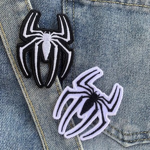 Può includere: Due toppe ricamate con un disegno di ragno. Una è nera con dettagli bianchi, l'altra bianca con dettagli neri. Le toppe sono posizionate su una giacca di jeans.