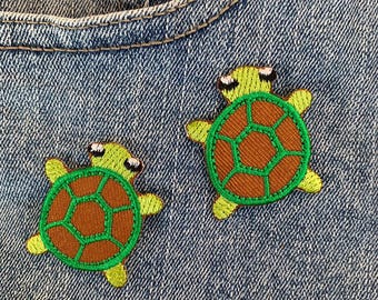 May include: Two green and brown embroidered turtle patches on a blue denim background.