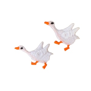 Op de afbeelding: Twee witte geborduurde ganzen-applicaties met oranje poten en zwarte ogen. De ganzen kijken in verschillende richtingen en hebben hun vleugels uitgespreid.