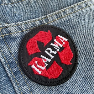 May include: A round black patch with the word "KARMA" in white lettering and red arrows. The patch is sewn onto a pair of blue denim jeans. The patch is a decorative item.