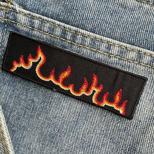 May include: A rectangular black embroidered patch featuring a stylised flame design in shades of red, orange, and yellow. The patch is stitched onto a light blue denim fabric with visible yellow topstitching along the seams of the garment.