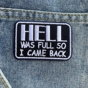 Könnte beinhalten: Ein rechteckiger schwarz-weißer Aufnäher mit dem Text "HELL WAS FULL SO I CAME BACK" in weißen Blockbuchstaben. Der Aufnäher ist auf einem blauen Denim-Stoff aufgenäht.