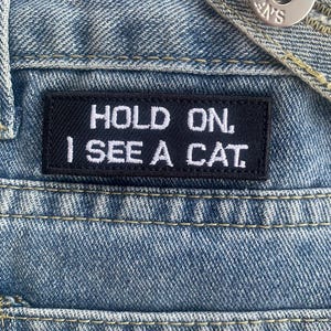 以下が含まれることがあります： 青いジーンズに、「Hold On, I See A Cat」と白く刺繍された黒いパッチが縫い付けられています。