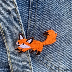 Peut inclure: Patch brodé de renard orange sur un fond en jean bleu.
