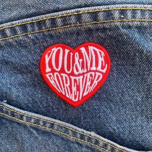 May include: Red heart-shaped patch with white embroidered text that reads "YOU & ME FOREVER" on a blue denim background.