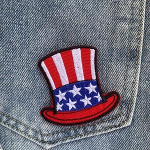 Puede incluir: Un parche patriótico con un diseño de sombrero del Tío Sam rojo, blanco y azul. El sombrero tiene rayas rojas y blancas, una banda azul con estrellas blancas y un borde rojo. El parche está cosido en un bolsillo de mezclilla.