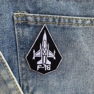 May include: Close-up of a denim pocket with a black and white F-16 patch. The patch is a diamond shape with a white jet plane design and the text "F-16" in white.