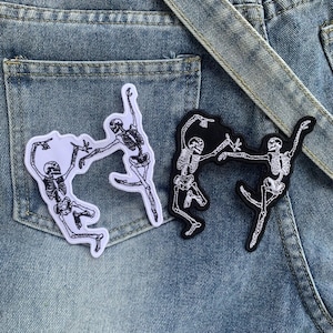 Könnte beinhalten: Zwei aufgestickte Aufnäher mit tanzenden Skeletten. Einer ist weiß mit schwarzen Details, der andere ist schwarz mit weißen Details. Die Aufnäher sind auf einem Jeanshintergrund platziert.