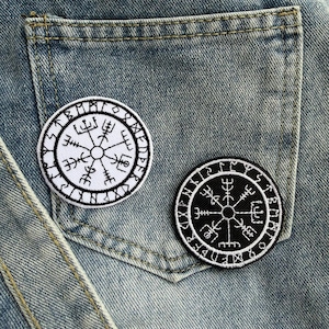 Könnte beinhalten: Zwei runde, gestickte Aufnäher mit einem Wikinger-Kompass-Design. Einer ist weiß mit schwarzen Nähten, der andere schwarz mit weißen Nähten. Sie befinden sich auf einem Jeans-Hintergrund.