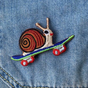 Puede incluir: Un parche bordado de un caracol montando un monopatín. El caracol es marrón con una concha roja y tiene un ojo negro. El monopatín es azul y verde con ruedas rojas.