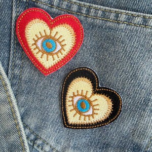 May include: Two heart-shaped embroidered patches with an eye design. One patch has a red border, the other a black border. Both have a cream-colored center with a blue eye and gold details, set against a denim background.