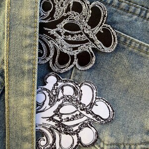 May include: Two embroidered octopus patches. One patch is black with white detailing, and the other is white with black detailing. The patches are placed on a blue denim background, with a denim seam visible on the left side.