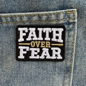 May include: A rectangular embroidered patch attached to light blue denim fabric. The patch features a black background with white stitched text that reads FAITH and FEAR, separated by a gold horizontal line with the word OVER in gold text.