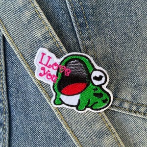 Può includere: Una toppa a forma di rana verde con bordo bianco e la scritta "I Love You" in rosa. La rana ha la bocca rossa e un occhio bianco. La toppa è su uno sfondo di denim.