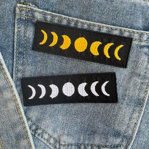 May include: Two rectangular patches with moon phase designs are displayed on a denim background. One patch features yellow moons on a black background, while the other has white moons on a black background. The patches are sewn onto the denim.