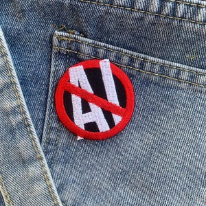 Puede incluir: Un c&iacute;rculo rojo con una l&iacute;nea diagonal que lo atraviesa, con las letras "AI" en blanco en su interior. El parche est&aacute; cosido en un bolsillo de mezclilla azul.