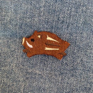 Peut inclure: Un écusson brodé marron représentant un sanglier courant vers la droite. Le sanglier a des rayures blanches et un œil noir.