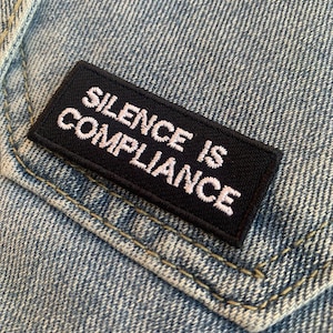 May include: Black embroidered patch with white text that reads "Silence is Compliance" on a blue denim background.
