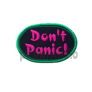 Puede incluir: Un parche ovalado negro con costuras verdes alrededor de los bordes. El texto "Don't Panic!" está bordado en rosa.