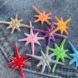 Può includere: Assortimento di toppe ricamate a forma di stella di vari colori. Le toppe sono di vari colori, tra cui rosso, arancione, giallo, verde, blu, viola, rosa e argento. Le toppe sono tutte della stessa dimensione e forma, e sono disposte in uno schema casuale su uno sfondo di denim blu.