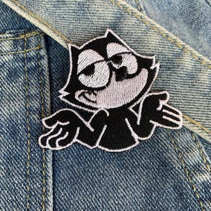 May include: Black and white embroidered iron-on patch of a cartoon cat with a mischievous expression. The cat is wearing a white collar and has large, round eyes.