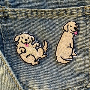 Op de afbeelding: Twee geborduurde patches van een golden retriever hond op een denim achtergrond. Eén patch toont de hond liggend, de andere de hond zittend. Beide patches hebben zwarte contouren en roze tongen.