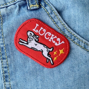 Könnte beinhalten: Roter gestickter Aufnäher mit einem weißen Kaninchen, das springt, und dem Wort "LUCKY" in weißen Buchstaben. Der Aufnäher ist auf einen blauen Jeansstoff genäht.