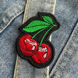 Op de afbeelding: Geborduurde patch met twee rode kersen met groene bladeren op een zwarte achtergrond. De patch is genaaid op een denim kledingstuk. De kersen hebben witte highlights en de bladeren zijn heldergroen.