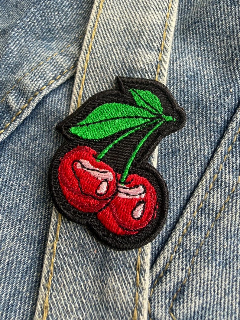 May include: Embroidered patch showing two red cherries with green leaves on a black backing. The patch is attached to denim. The cherries have white highlights, and the leaves are a bright green.