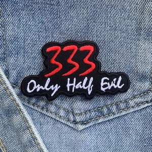 Könnte beinhalten: Ein schwarzer, gestickter Aufnäher mit der roten Zahl "333" und den weißen Worten "Only Half Evil". Der Aufnäher ist auf einer Jeansjackentasche angebracht.